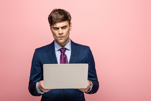 thoughtful businessman in suit standing with laptop on pink background - Photo, Image
