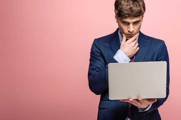 serious businessman in suit using laptop and holding hand near face on pink background - Fotoğraf, Görsel