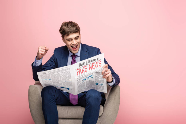 vrolijke zakenman toont winnaar gebaar terwijl zittend in fauteuil en lezing krant met nep nieuws op roze achtergrond - Foto, afbeelding
