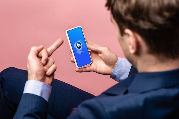 KYIV, UKRAINE - MAY 16, 2019: cropped view of businessman using smartphone with shazam app and showing middle finger, isolated on pink - Foto, immagini