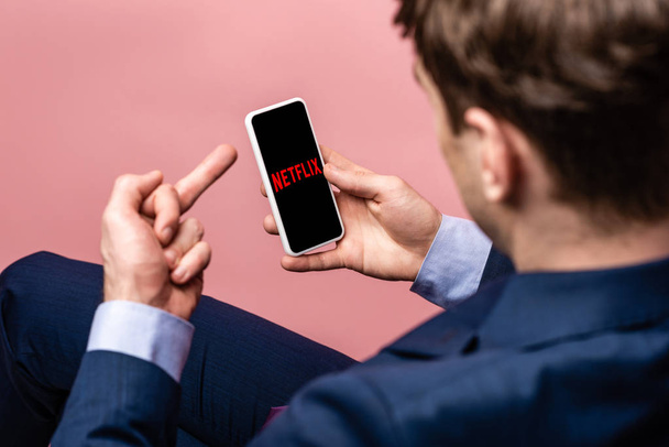 KYIV, UKRAINE - MAY 16, 2019: cropped view of businessman using smartphone with netflix app and showing middle finger, isolated on pink - Фото, зображення