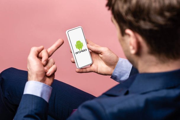 KYIV, UKRAINE - MAY 16, 2019: cropped view of businessman using smartphone with android app and showing middle finger, isolated on pink - 写真・画像