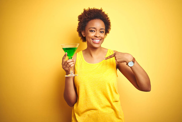 Giovane donna afro-americana con capelli afro bere un cocktail su sfondo giallo isolato con faccia a sorpresa puntando il dito verso se stesso
 - Foto, immagini