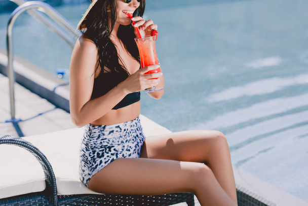 cropped view of smiling beautiful brunette young woman in swimsuit drinking cocktail while sitting on sunbed - Foto, afbeelding