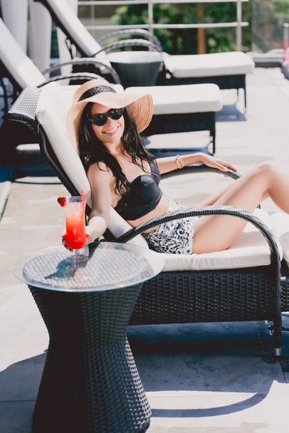 heureuse belle brune jeune femme en maillot de bain, chapeau de paille et lunettes de soleil bronzant et tenant cocktail fraise glacée sur la table
 - Photo, image