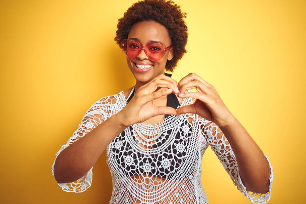 Afro saçlı, bikini ve kalp şeklinde güneş gözlüğü takan, kalp sembolü ve elleri ile gülümseyen Afro Amerikalı genç bir kadın. Romantik konsept. - Fotoğraf, Görsel