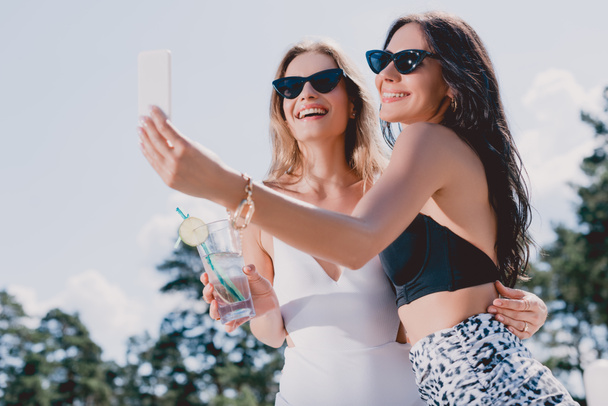 low angle view of happy brunette and blonde women in swimsuits taking selfie - Photo, Image