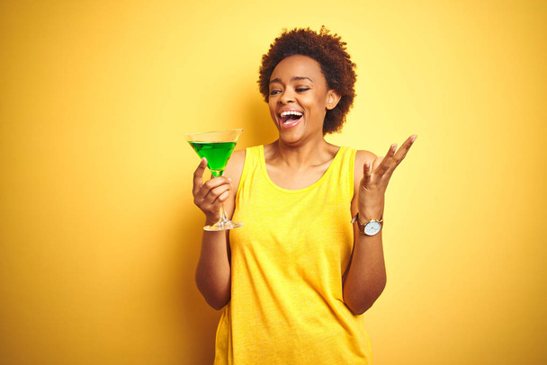 Молода афроамериканська жінка з волоссям афроамериканського кольору п'є коктейль над жовтим оточенням дуже щаслива і схвильована, переможний вираз, що прославляє перемогу з великою посмішкою і піднятими руками. - Фото, зображення