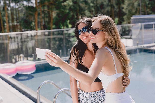 boldog szőke és barna nők napszemüveg és fürdőruhák vesz selfie közelében medence  - Fotó, kép