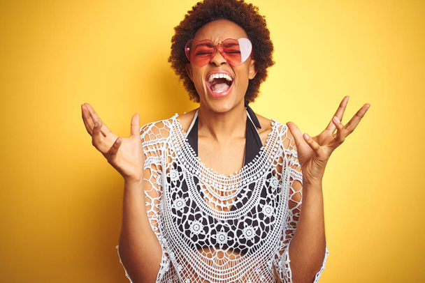Jeune femme afro-américaine aux cheveux afro portant un bikini et des lunettes de soleil en forme de cœur célébrant fou et fou de succès avec les bras levés et les yeux fermés hurlant excités. Concept gagnant
 - Photo, image