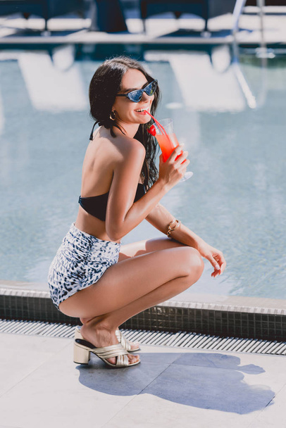 šťastná sexy brunetka v plavkách a slunečních brýlích, kde sedí nedaleko bazénu a pije koktejl  - Fotografie, Obrázek