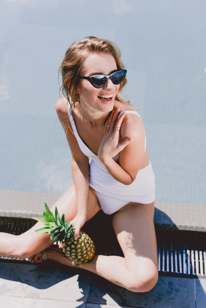 šťastná blondýnka v brýlích a plavkách s ananasem poblíž bazénu - Fotografie, Obrázek