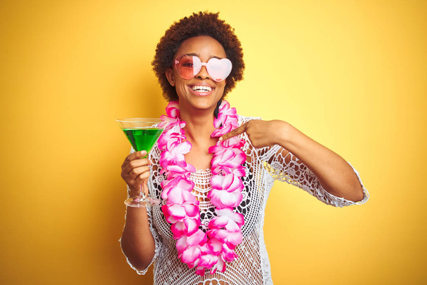Молода афроамериканська жінка з волоссям афроамериканця, одягнена в квіткове леї і п'є коктейль з несподіваним обличчям, вказуючи пальцем на себе. - Фото, зображення