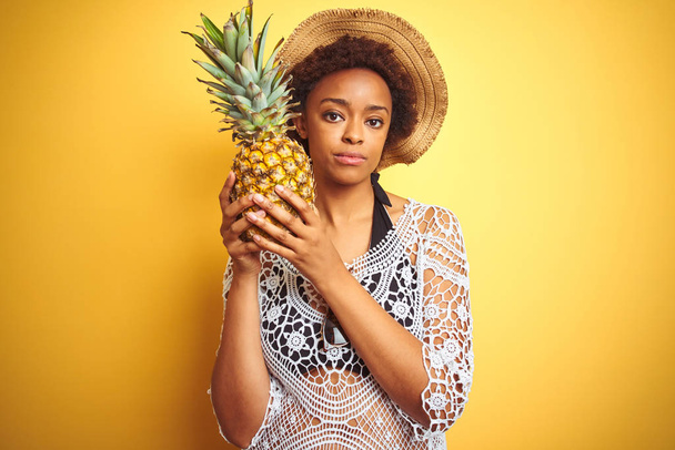 Joven mujer afroamericana con cabello afro sosteniendo piña sobre fondo aislado amarillo con una expresión segura en cara inteligente pensando seriamente
 - Foto, Imagen