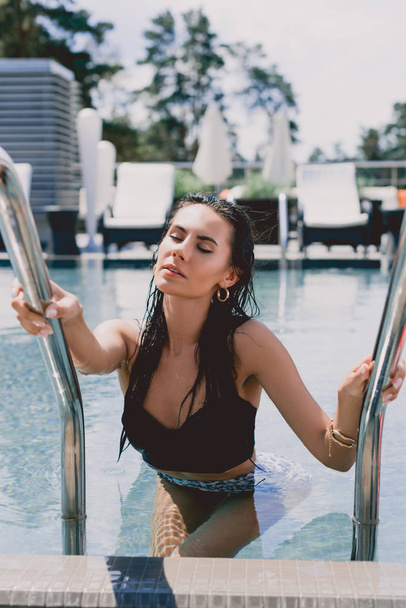wet brunette woman with closed eyes and hands on metal handrails getting out from water in swimming pool - Фото, зображення