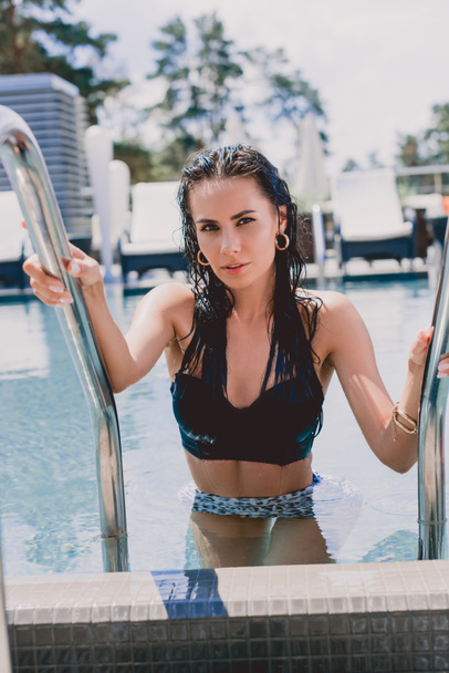 wet brunette woman with hands on metal handrails getting out from water in swimming pool and looking at camera - Фото, изображение