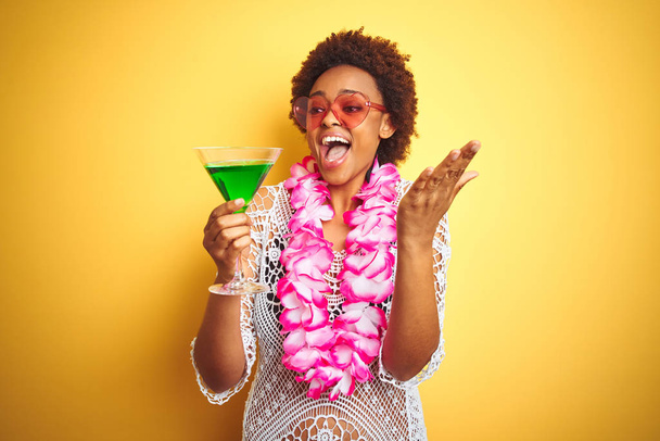Jeune femme afro-américaine aux cheveux afro portant une fleur hawaïenne lei et buvant un cocktail très heureux et excité, expression gagnante célébrant la victoire en criant avec un grand sourire et les mains levées
 - Photo, image