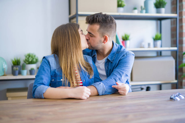 Νεαρό όμορφο ζευγάρι που κάθεται στο τραπέζι στο σπίτι, αγκαλιάζοντας την αγάπη πολύ χαρούμενη για τη μετακόμιση σε νέο σπίτι με χαρτοκιβώτια πίσω τους - Φωτογραφία, εικόνα