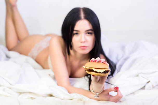 Eating junk food in bed. Girl in sexy lingerie eat burger. Sexual appetite. Food delivery service. Seductive sexy woman relax on bed. Diet concept. Fast food. Seductive woman hungry. Lazy day food - Foto, Bild