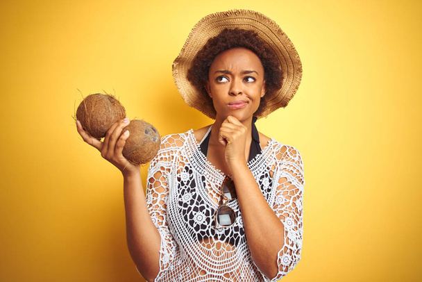 Jovem mulher afro-americana com cabelo afro segurando coco sobre fundo isolado amarelo rosto sério pensando em questão, ideia muito confusa
 - Foto, Imagem