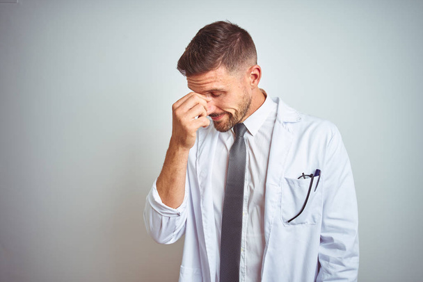 Young handsome doctor man wearing white profressional coat over isolated background tired rubbing nose and eyes feeling fatigue and headache. Stress and frustration concept. - Photo, Image