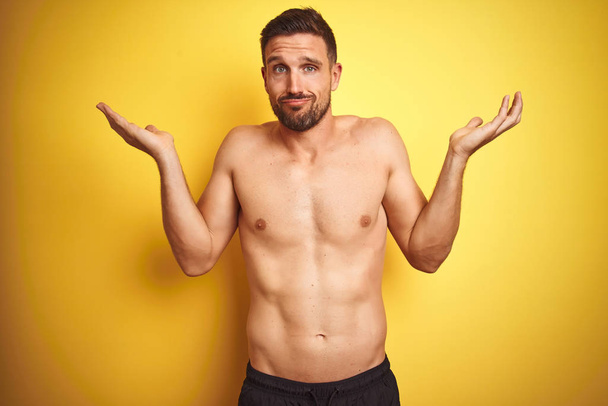 Young handsome shirtless man over isolated yellow background clueless and confused expression with arms and hands raised. Doubt concept. - Photo, Image