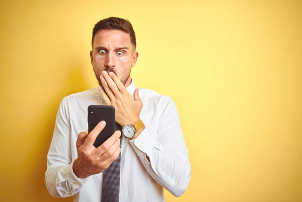 Młody przystojny biznesmen korzystający ze smartfona nad żółtym, odizolowanym tłem usta z ręką zaszokowany wstydem za pomyłkę, wyrażeniem strachu, przerażony w ciszy, sekretny koncept - Zdjęcie, obraz