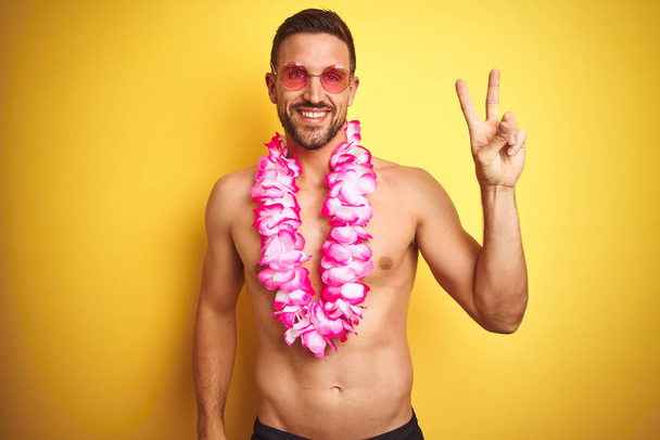 Young handsome shirtless man wearing sunglasses and pink hawaiian lei over yellow background smiling with happy face winking at the camera doing victory sign. Number two. - Photo, Image