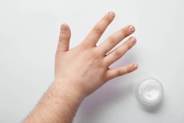 vista cortada do homem segurando a mão sobre a superfície branca com creme cosmético no frasco
 - Foto, Imagem