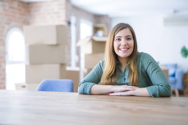 Mujer joven sentada en la mesa con cajas de cartón detrás de su mudanza a un nuevo hogar con una sonrisa feliz y fresca en la cara. Persona afortunada
. - Foto, Imagen