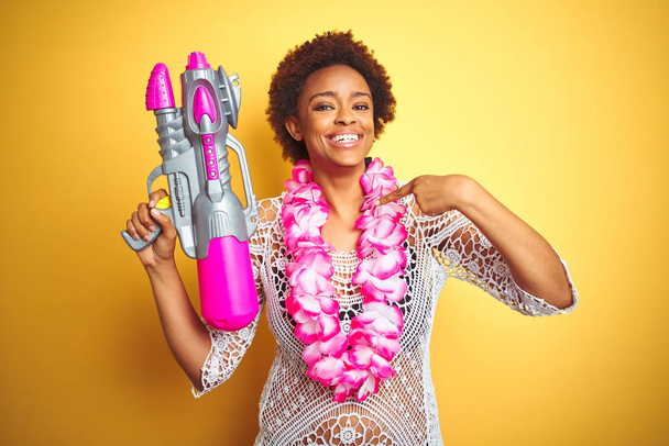 Joven mujer afroamericana con pelo afro vistiendo flor hawaiana lei y pistola de agua con la cara sorpresa señalando el dedo a sí mismo
 - Foto, Imagen