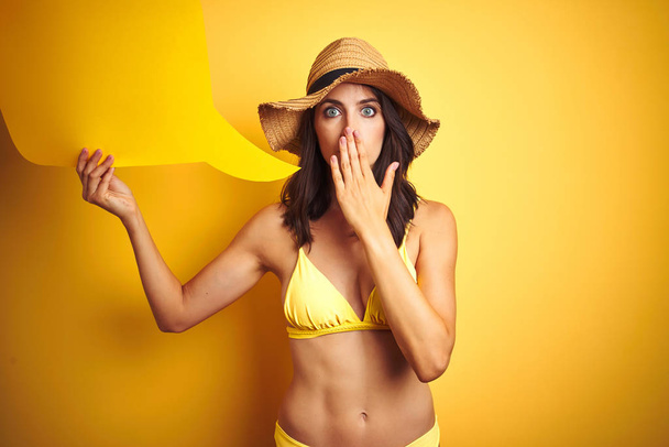 Beautiful woman wearing yellow bikini and holding talking balloon over isolated yellow background cover mouth with hand shocked with shame for mistake, expression of fear, scared in silence, secret concept - Photo, Image