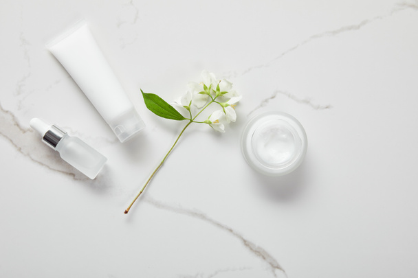 top view of cosmetic glass bottle, jar with cream, moisturizer tube and jasmine on white surface - Photo, Image