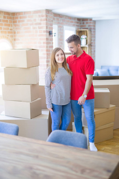 Young beautiful couple in love around cardboard boxes moving to new home, smiling very happy and cheerful for new apartment - Foto, Imagem