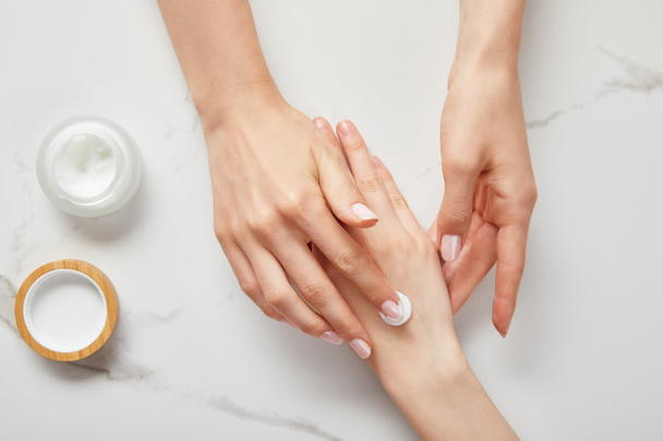 cropped view of woman applying cream on hand, using white moisturizer from jar on white surface - Fotó, kép