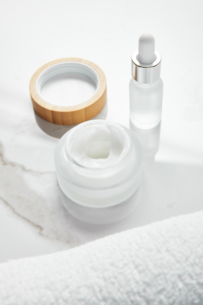ανοιχτό βάζο με καλλυντική κρέμα και γυάλινο μπουκάλι κοντά πετσέτα σε λευκή επιφάνεια - Φωτογραφία, εικόνα