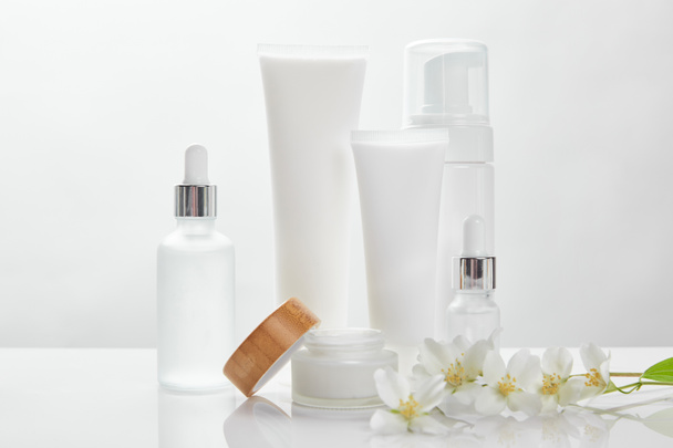 jasmine flowers on white surface near glass bottles, cream in tubes, jar and cosmetic dispenser - Zdjęcie, obraz