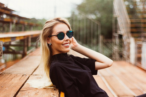 Portrait of a beautiful young woman. She is wearing sunglasses and smiling happily sitting on the bleachers. People, facial expressions and positive emotions. Lifestyle - Foto, Imagen