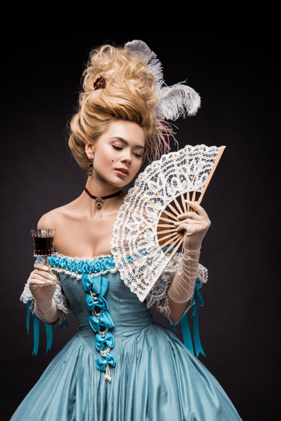 attractive victorian woman holding fan and wine glass while standing on black  - Valokuva, kuva