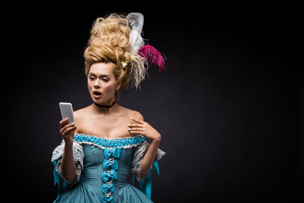 shocked young victorian woman in blue dress looking at smartphone on black  - Zdjęcie, obraz