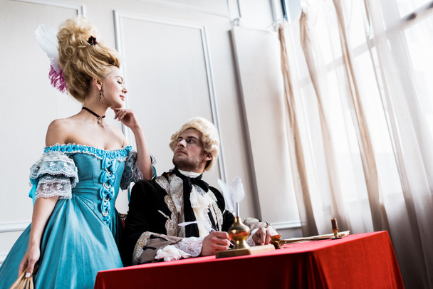 low angle view of victorian girl in wig standing near handsome pompous man holding feather near paper  - Foto, Imagem