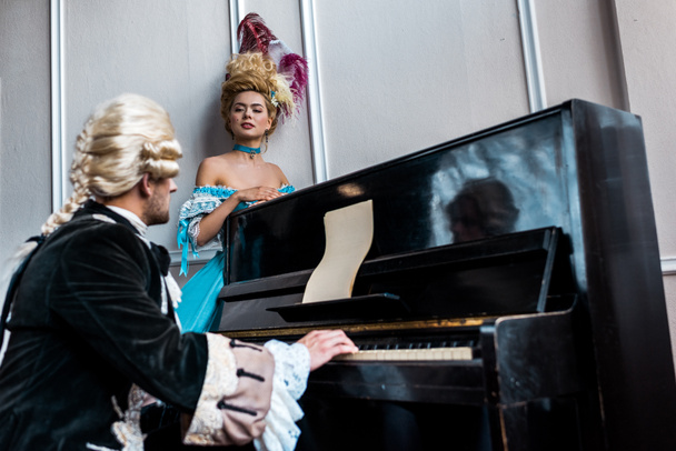 επιλεκτική εστίαση της Βικτωριανής γυναίκας κοιτάζοντας τον άνθρωπο με περούκα παίζοντας πιάνο  - Φωτογραφία, εικόνα