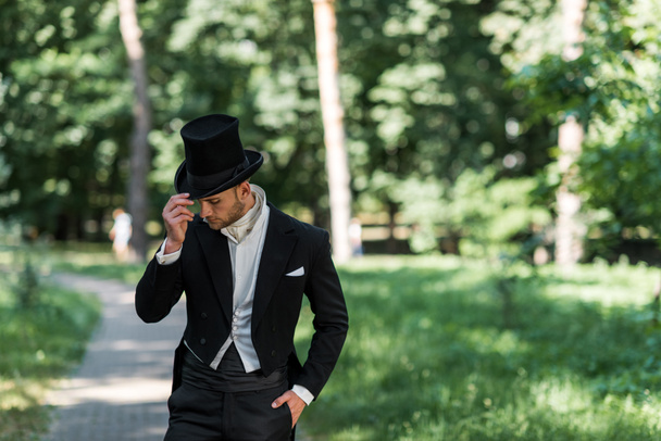 handsome young victorian man touching hat and standing with hand in pocket outside  - Фото, изображение