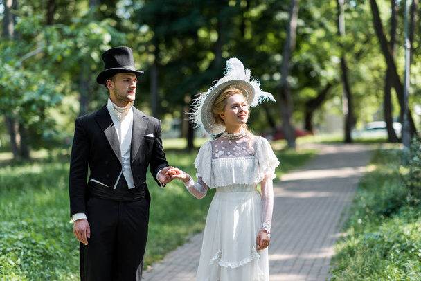 victorian man and woman in hats holding hands while standing outside near green trees  - Photo, Image