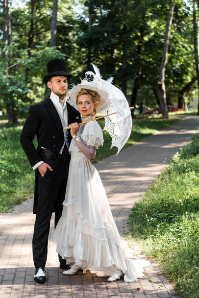 傘を持つ女性の近くにポケットに手を持って立っているハンサムなビクトリア朝の男  - 写真・画像