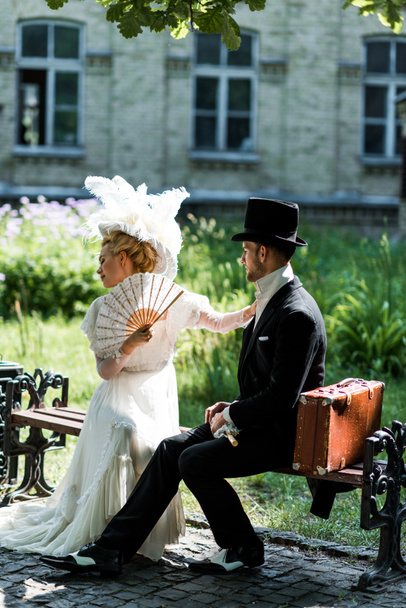 attractive victorian woman holding fan while sitting with handsome man on bench  - Foto, afbeelding