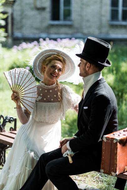 glückliche viktorianische Frau hält Ventilator, während sie mit hübschem Mann auf Bank sitzt  - Foto, Bild