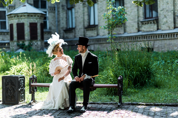 fröhliche viktorianische Frau hält einen Fächer, während sie mit einem jungen Mann auf einer Bank sitzt  - Foto, Bild