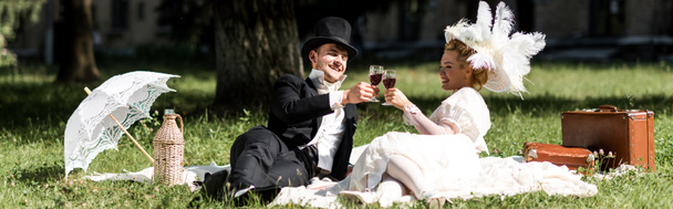 panoramic shot of happy man and woman siting on blanket and clinking wine glasses  - Фото, изображение