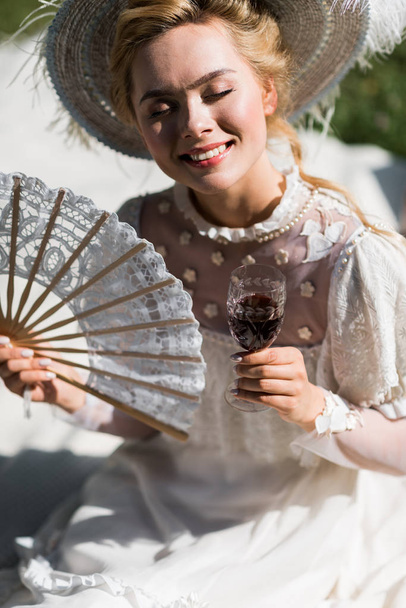feliz joven victoriana con los ojos cerrados sosteniendo ventilador y copa de vino
  - Foto, imagen